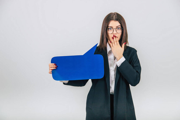 Portrait of an excited young business woman holding empty square blue speech bubble, covering her mouth with hand, surprised face. - Fotografie, Obrázek