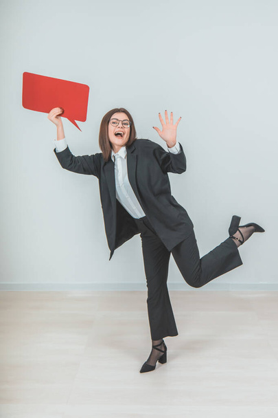 Full-length excited young business woman standing on one leg, raising up empty square red speech bubble, looking at the camera, smiling. - Foto, afbeelding