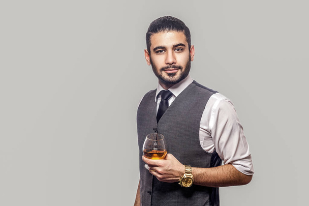 handsome bearded man in white shirt and waistcoat holding glass of whiskey and looking at camera with serious face on gray background - Foto, afbeelding