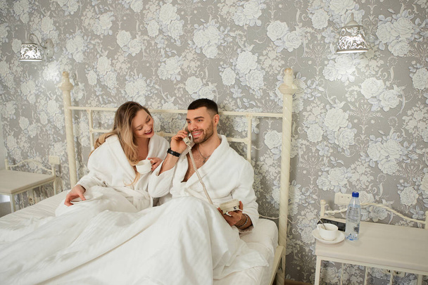 Joyeux jeune couple en manteaux blancs, ils se réveillent et commandent le petit déjeuner dans la chambre par téléphone
 - Photo, image