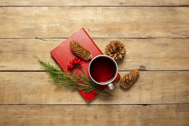Book, metal cup with hot tea and cones on a wooden background. The concept of the onset of autumn, winter, vacation in a country house, camping trip. Flat lay, top view - Photo, Image