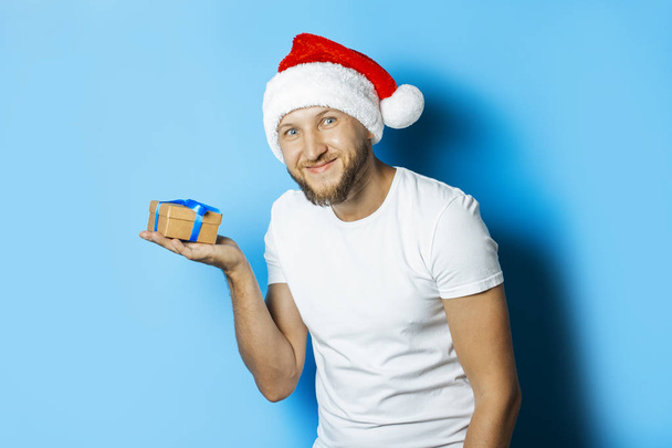 Mężczyzna w kapeluszu Świętego Mikołaja trzyma prezent na niebieskim tle. - Zdjęcie, obraz