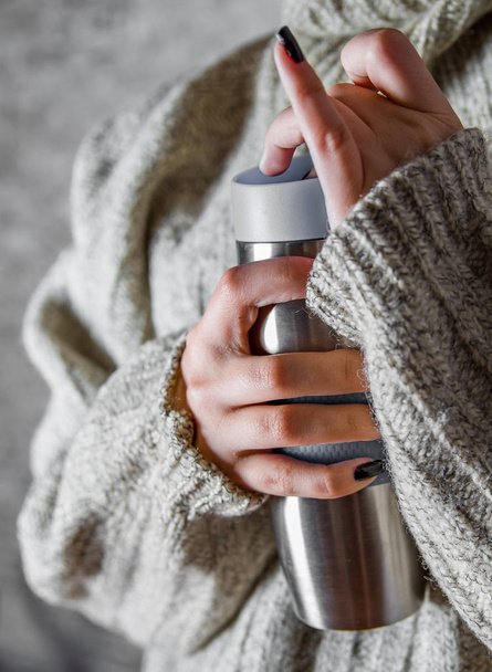 donna in un maglione tiene un termomug con una bevanda calda tra le mani su sfondo grigio parete
 - Foto, immagini