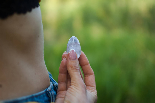 Вид сбоку на взрослую женщину, держащую прозрачное фиолетовое яйцо аметист-йони для фьюмфита, имбилдинга или медитации на открытом воздухе над её обнаженным животом. Концепция женского здоровья
 - Фото, изображение