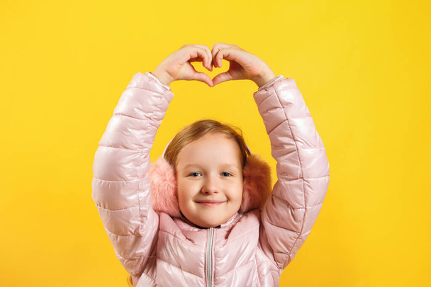 Cheerful little girl shows a heart with her fingers over her head. A child in a jacket and warm ear muffs on a yellow background. The concept of winter, christmas, new year - Photo, image