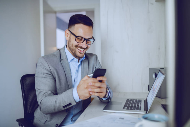 Lachen aantrekkelijke stijlvolle blanke zakenman in grijs pak en met bril met behulp van een smartphone voor sms-bericht tijdens het zitten in een modern kantoor. Op het bureau ligt een laptop. - Foto, afbeelding