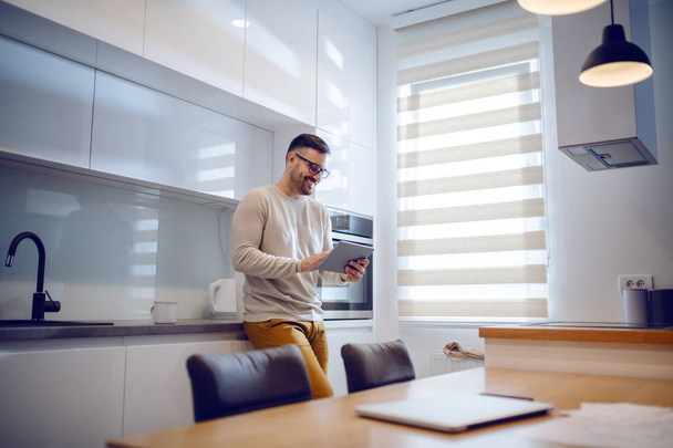 Positive beautiful Caucasian man dressed casual standing in modern kitchen and surfing on internet over tablet. - Fotografie, Obrázek