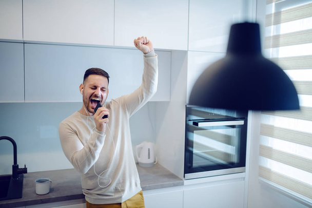Playful handsome caucasian man dressed casual listening his favorite music over smart phone and pretending to sing on microphone. Modern domestic kitchen interior. - Фото, зображення