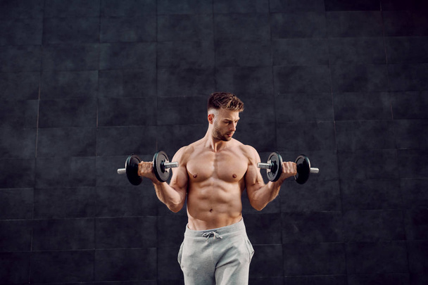 Handsome muscular caucasian blond shirtless man lifting dumbbells while standing in front of dark background. - Foto, immagini