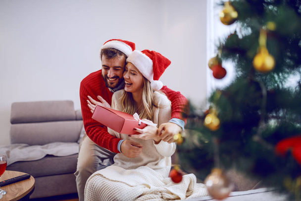 Surprised smiling caucasian woman sitting on sofa in living room and holding gift. Man hugging woman. Both having santa hats on heads. In foreground is christmas tree. Living room interior. - Φωτογραφία, εικόνα