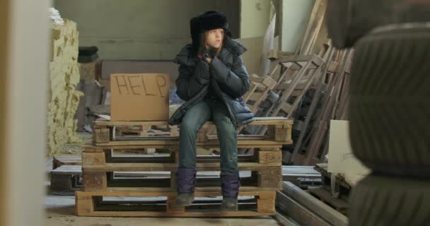 Little Syrian refugee in dirty warm clothes sitting on pallets at the construction site with Help cardboard. Lonely homeless child waiting for a handout. - Séquence, vidéo