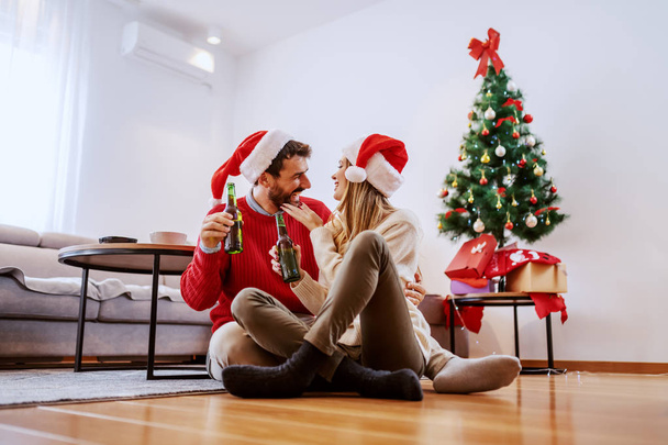 entzückendes, gutaussehendes kaukasisches Paar mit Weihnachtsmützen auf dem Kopf, das auf dem Boden im Wohnzimmer sitzt, kuschelt und Bier in der Hand hält. Im Hintergrund ein Weihnachtsbaum mit Geschenken. - Foto, Bild
