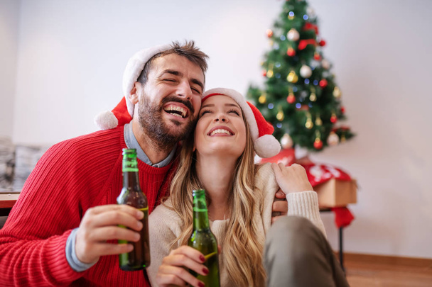 entzückendes, gutaussehendes kaukasisches Paar mit Weihnachtsmannmützen auf dem Kopf, das auf dem Boden im Wohnzimmer sitzt, sich umarmt und Bier in der Hand hält. Im Hintergrund ein Weihnachtsbaum mit Geschenken. - Foto, Bild