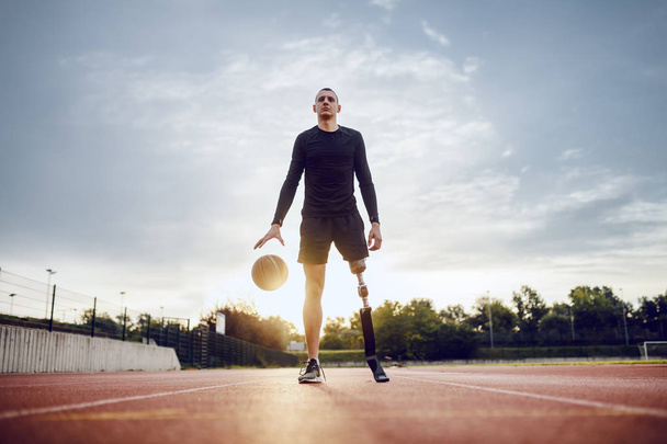 Toute la longueur de l'homme handicapé caucasien sportif en vêtements de sport et jambe artificielle dribble la balle tout en se tenant debout sur l'hippodrome
. - Photo, image