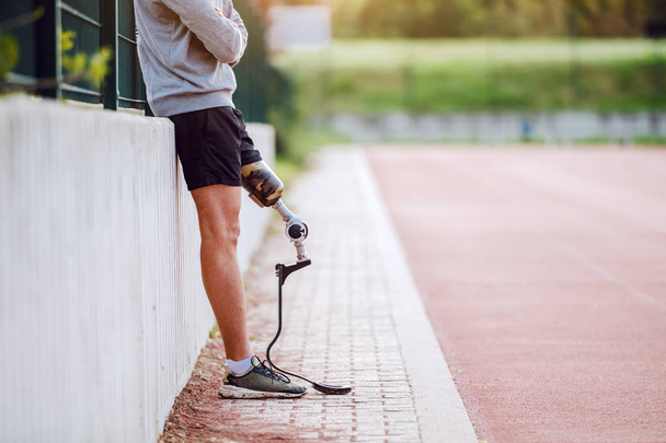Κόψτε την εικόνα του όμορφος αθλητικός ανάπηρος άνδρας σε αθλητικά ρούχα και με τεχνητό πόδι ακουμπά στο φράχτη, ενώ κρατώντας τα χέρια σταυρωμένα. - Φωτογραφία, εικόνα