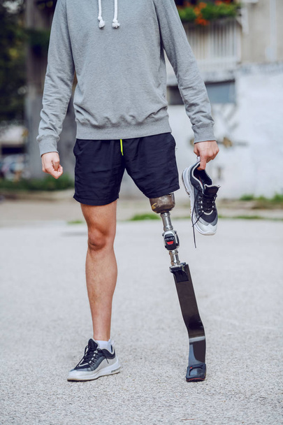 Cortar imagen de hombre deportivo discapacitado de pie al aire libre con zapatillas de deporte en la mano
. - Foto, Imagen