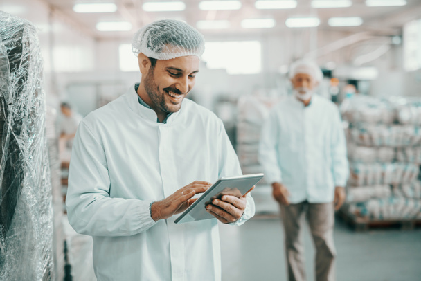 Sonriente supervisor caucásico en uniforme estéril de pie en la fábrica de alimentos con la tableta en las manos y el control de la calidad de las mercancías
. - Foto, Imagen