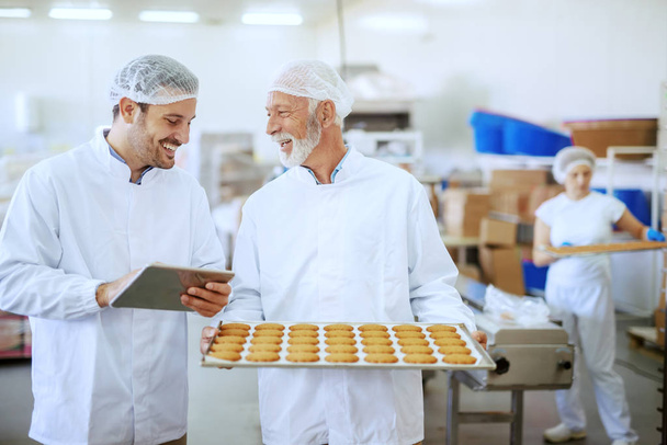 Senior adult employee holding tray with fresh cookies while supervisor evaluating quality and holding tablet. Both are dressed in sterile white uniforms and having hairnets. Food plant interior. - Photo, Image
