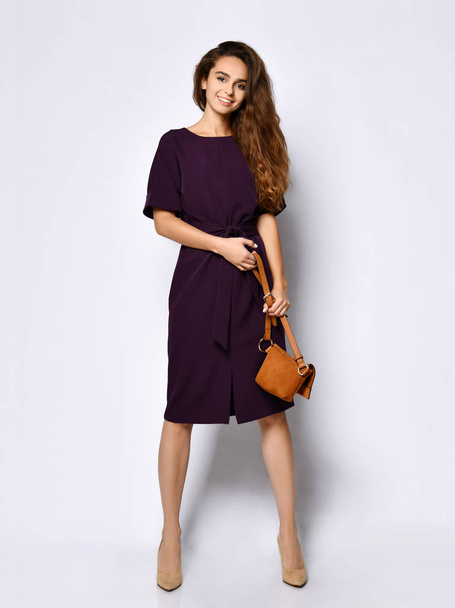 slender curly female model in a purple dark long dress and a small brown handbag, in full growth. - 写真・画像