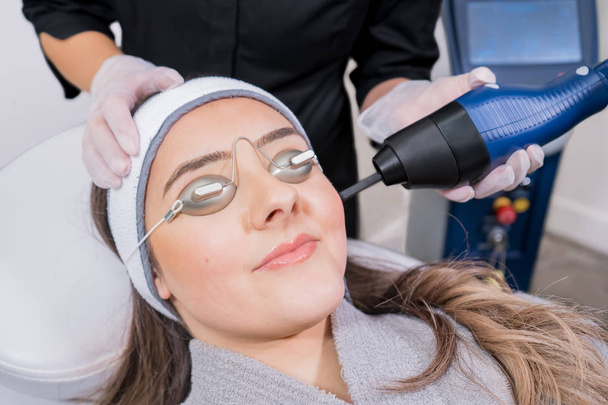 Láser ablativo fraccional de CO2 que se utiliza para el rejuvenecimiento de la piel (rejuvenecimiento de la piel) como un procedimiento cosmético médico en una clínica de láser de belleza. Paciente femenina con gafas, con técnica láser de belleza
 - Foto, imagen