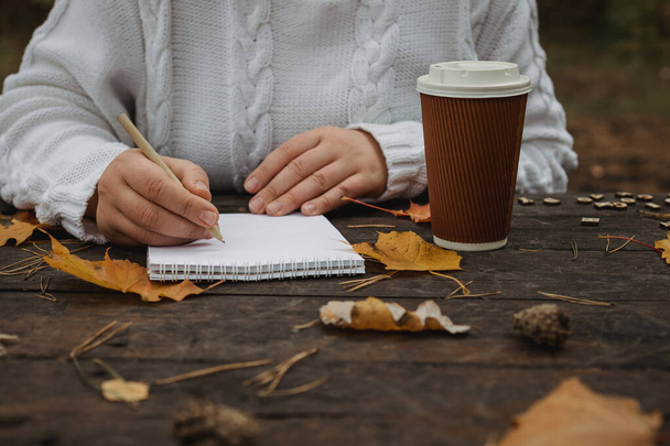 ノートとペンと秋の葉を持つ木製のテーブルでコーヒーとスカーフのカップを持つ人間の手。木製のテーブルでコーヒーとスカーフのカップを持つ人間の手 - 写真・画像