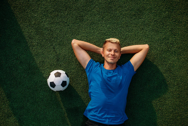 Male soccer player with ball lying on the grass on the field, top view. Footballer on outdoor stadium, workout before game, football training - Photo, Image