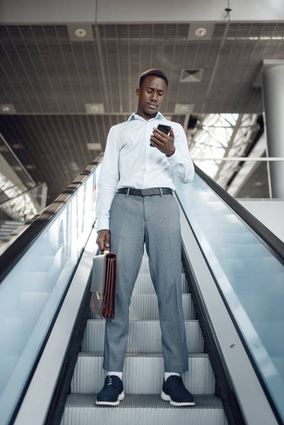 Uomo d'affari africano con valigetta che parla per telefono sulla scala mobile nel centro commerciale
.  - Foto, immagini
