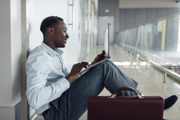Afrikanischer Geschäftsmann sitzt mit Aktentasche und Laptop auf dem Fußboden im Büroflur.  - Foto, Bild