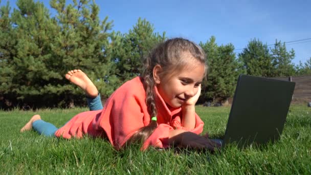 A girl of seven years old works on a laptop on the green grass. Distance learning. Girl with a laptop on a background of pines. Girl with a computer on the street. A child on the grass plays computer games. Remote education. - Video