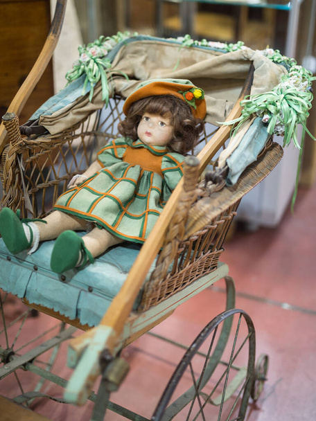 Boneca antiga com vestido verde e laranja descansando em um carrinho de madeira e vime
 - Foto, Imagem