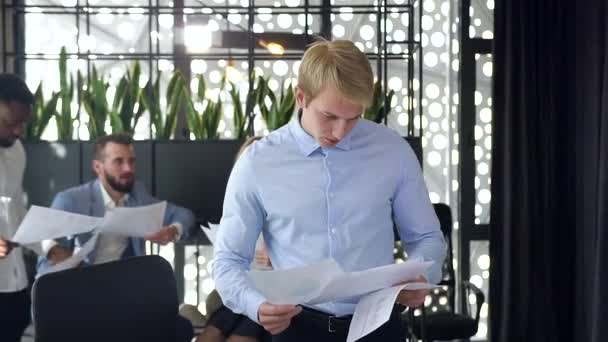 Attractive portrait of serious young blond businessman coming near to his workplace and starts to work with computer on the coworkers background - Felvétel, videó