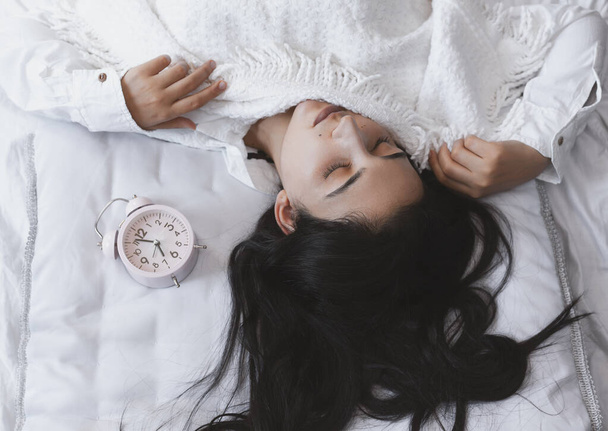 Beautiful black-haired woman sleeping in cozy bed. The alarm clock set at 7 a.m. morning. Female is going to wake up at 7 o'clock early morning. Top view - Photo, Image