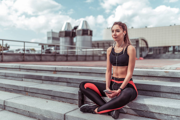 Athlete woman resting in summer on steps city. Rest after workout with fitness and outdoor workout. In hands phone, listens music online application on Internet. Free space for text. - Valokuva, kuva