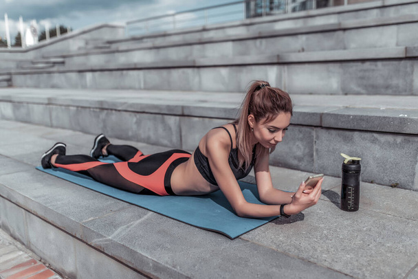 Athlete woman resting in summer in city on yoga mat. Rest after a workout with fitness outdoor workout. In hands of phone, an online application on the Internet. - Fotografie, Obrázek