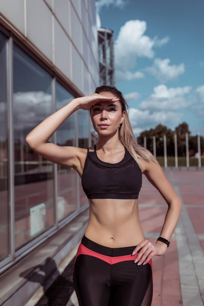 athlete girl in summer in city looks into distance, covering her hand from bright sun. Workout before morning jogging, fitness outdoor workout. Tanned slim figure, motivation woman lifestyle. - Foto, Imagem
