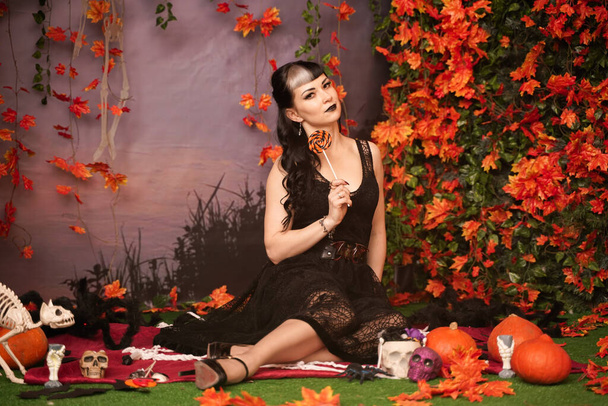Chica de halloween de moda delgada con pelo negro en encaje gótico pin up vestido posando en el fondo de otoño con hojas de otoño
 - Foto, Imagen
