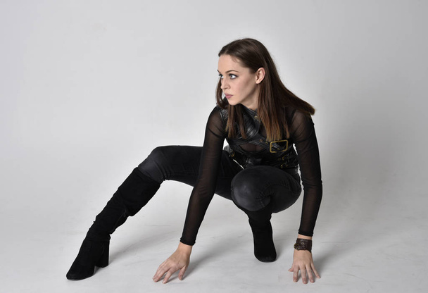 full length portrait of a pretty brunette woman wearing black leather fantasy costume. Crouching pose on a studio background. - Photo, Image