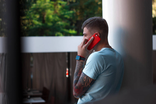Handsome guy is talking on the mobile phone outside. Modern stylish young man with a tattoo and smart watch on his arm is answering the phone. His smartphone is in a red case. Back view - Fotoğraf, Görsel