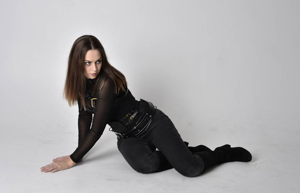 full length portrait of a pretty brunette woman wearing black leather fantasy costume. Crouching pose on a studio background. - 写真・画像