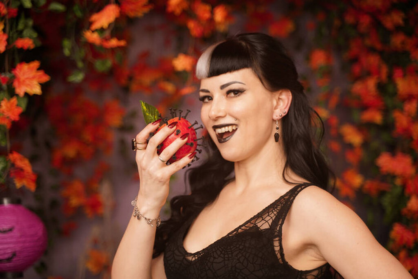 beautiful halloween girl in black pin up clothes with dead red apple in the hand on autumn leaves background alone - Photo, Image