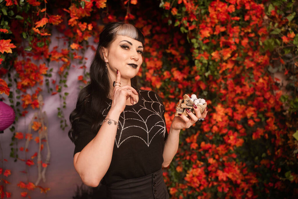 glückliche junge Frau mit schwarzem Haar, die Schokoladenaugen Bonbons aus Totenkopf hält und isst. Studioporträt über Herbst Blätter Hintergrund - Foto, Bild