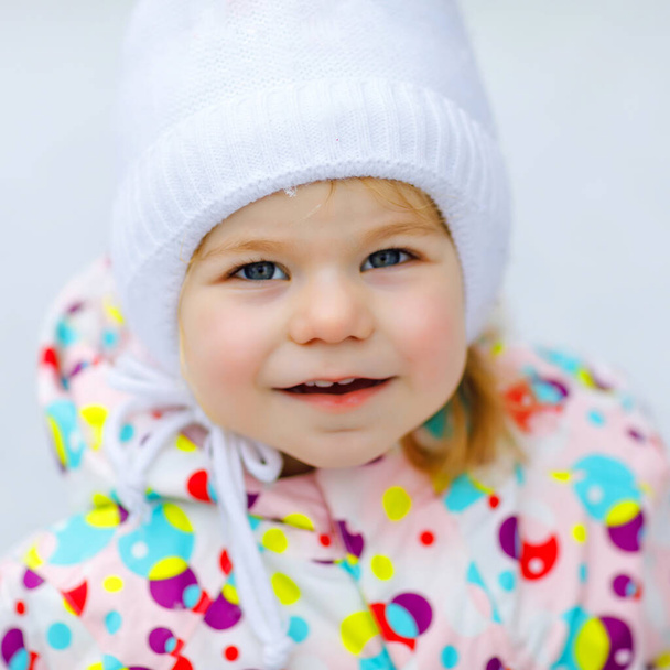 Portrait of little toddler girl walking outdoors in winter. Cute toddler eating sweet lollypop candy. Child having fun on cold snow day. Wearing warm baby colorful clothes and hat with bobbles. - Photo, Image