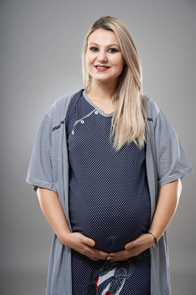 Young pregnant woman in pajamas, studio portrait on gray background  - Fotografie, Obrázek