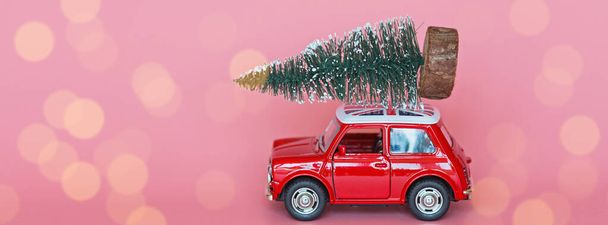 Czerwony samochodzik z choinką na dachu na różowym papierowym tle. Dostawa zimowa, Boże Narodzenie, Szczęśliwego Nowego Roku 2020 koncepcja uroczystości. kartka okolicznościowa, makieta, przestrzeń do kopiowania, miejsce na tekst, szablon - Zdjęcie, obraz