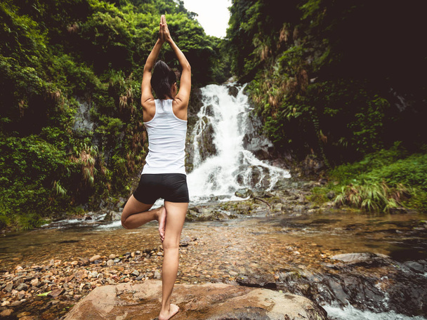 Fit young woman practicing yoga near waterfall in forest - Photo, Image