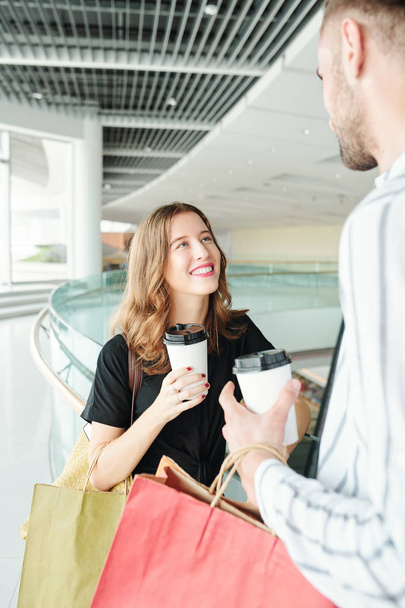 radosna ładna młoda kobieta patrząc na jej wysoki chłopak podczas picia kawy i dyskusji z jego sprzedaży i zakupów - Zdjęcie, obraz