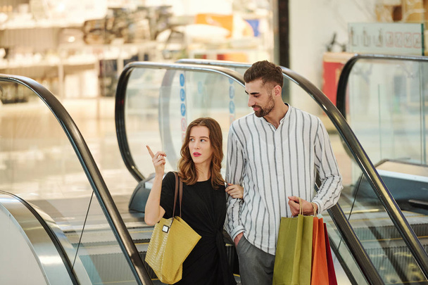 Pretty young woman pointing at boutique in shopping mall when standing on escalator with her husband - Zdjęcie, obraz