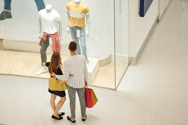 Abrazar a pareja joven con bolsas de compras mirando ropa masculina en el escaparate de la boutique de moda
 - Foto, Imagen