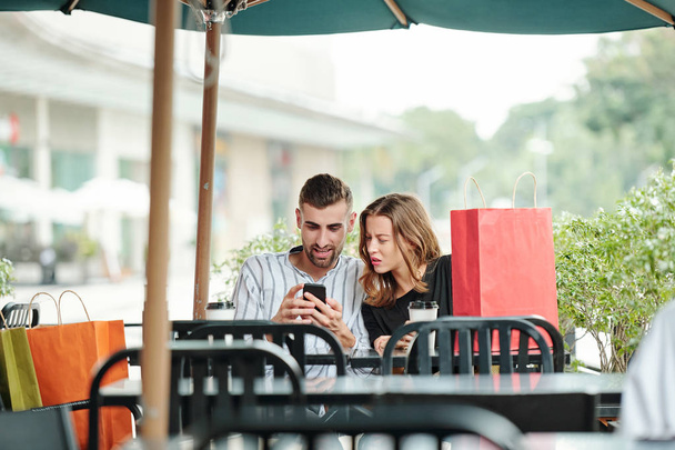 Frowning young woman looking at screen of smartphone in hands of her boyfriend when they are sitting at cafe table after shopping - Photo, Image