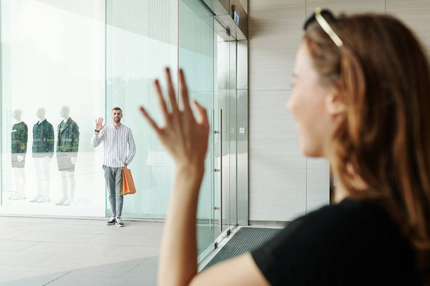 Mujer joven de pie en la ventana del centro comercial y saludando a su alegre novio de pie fuera
 - Foto, Imagen